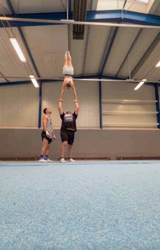 Trainingsvideo: Flyerin im Handstand auf den Händen ihrer Base, daneben ein Spotter